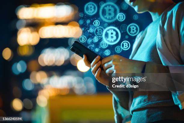 mid-section of young asian woman using smartphone in city at night, against illuminated street lights bokeh, working with bitcoin technologies, investing or trading bitcoin on cryptocurrency. business on the go - bitcóin fotografías e imágenes de stock