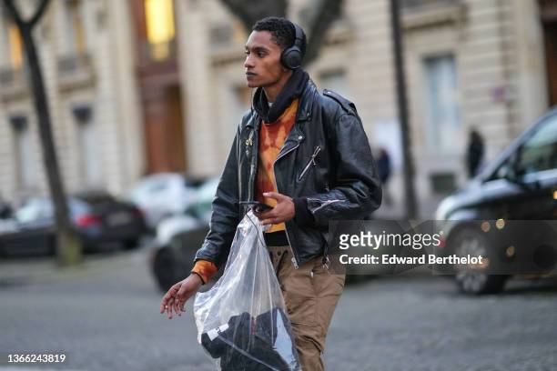 Model wears a white high neck t-shirt, a black hoodie sweater, an orange and brown tie and dye sweater, a black shiny leather jacket, a transparent...