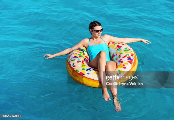 portrait of modern  beautiful young woman in sunglasses and turquoise color swimming suit swims in the sea with colorful inflatable ring. - float stockfoto's en -beelden