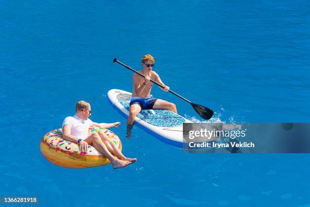 two young  attractive men in sunglasses swim in the sea with colorful inflatable ring and  stand up paddle sup board with oars. - surf tube stock pictures, royalty-free photos & images