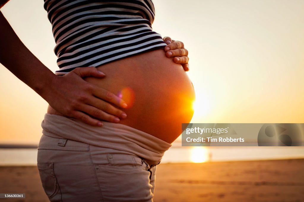 Pregnant woman holding bump on beach at sunset.