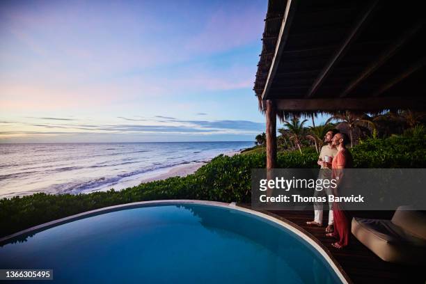 wide shot of gay couple standing poolside watching sunrise at luxury tropical villa overlooking beach and ocean - fabulous full lengths stockfoto's en -beelden