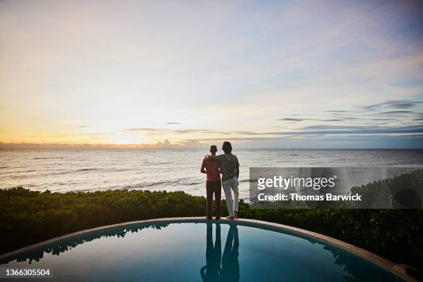 wide shot of embracing gay couple watching sunrise while standing at edge of pool at luxury tropical villa overlooking ocean - choicepix stock pictures, royalty-free photos & images
