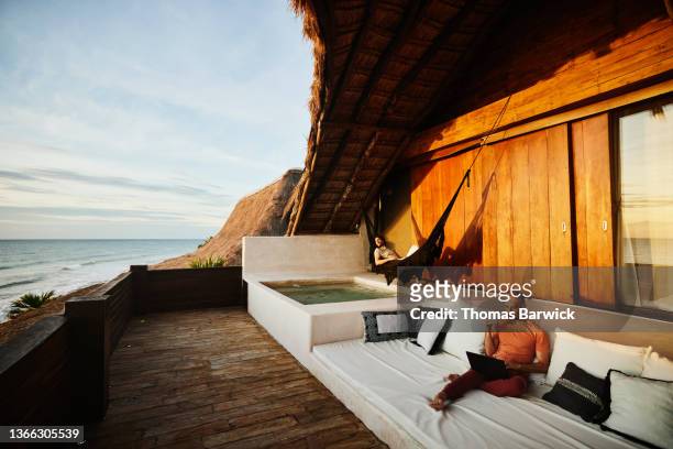 Wide shot of man drinking coffee and working on laptop while partner relaxes in hammock on deck of luxury tropical beach villa at sunrise