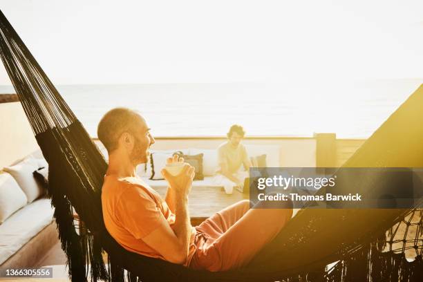 medium wide shot of man sitting in hammock watching sunrise while partner works on laptop from deck of luxury tropical beach villa - sea cup stock pictures, royalty-free photos & images