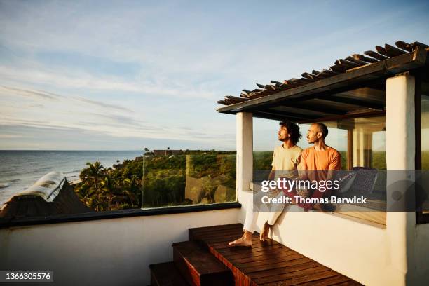 wide shot of gay couple sitting on rooftop deck of luxury tropical beachfront villa while drinking coffee and watching sunrise - coffee culture stock-fotos und bilder