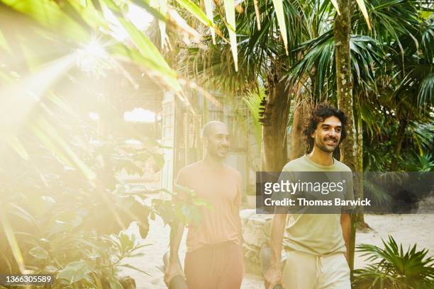 Medium wide shot of smiling gay couple walking through jungle at tropical resort on way to yoga class