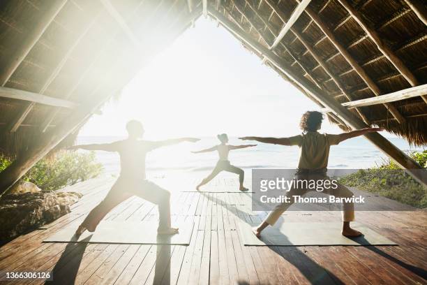 Wide shot of gay couple taking sunrise yoga class in beachfront pavilion at luxury tropical resort