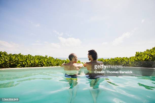 Wide shot rear view of gay couple relaxing in pool at luxury tropical beachfront villa looking at view