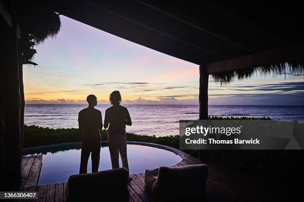 wide shot rear view of gay couple standing poolside watching sunrise at luxury tropical villa overlooking beach and ocean - poolside stock pictures, royalty-free photos & images