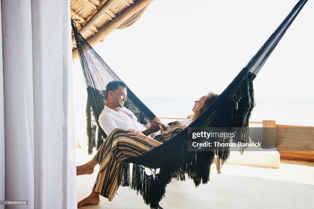 Medium wide shot of smiling couple holding hands while sitting in hammock on deck of luxury tropical villa
