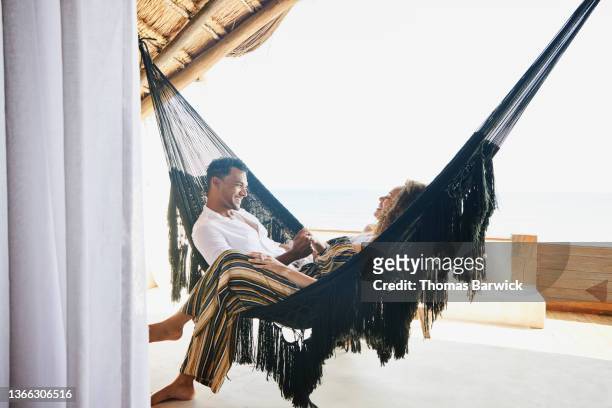 medium wide shot of smiling couple holding hands while sitting in hammock on deck of luxury tropical villa - bonding stock pictures, royalty-free photos & images