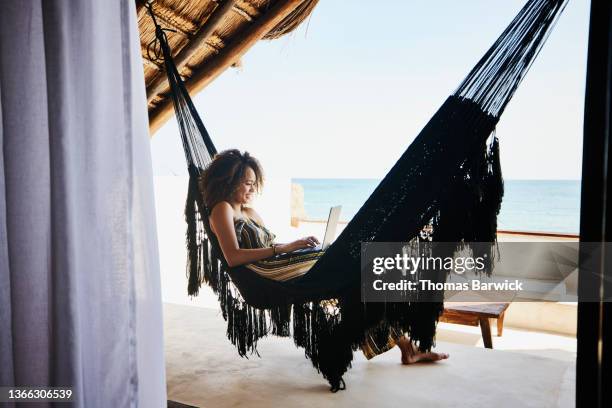 wide shot of woman working on laptop while relaxing in hammock on deck of luxury tropical villa overlooking ocean - luxury holiday stock pictures, royalty-free photos & images