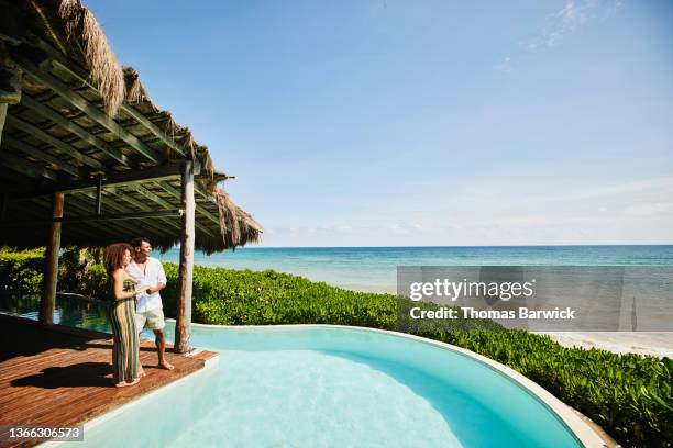 wide shot of couple standing poolside at luxury tropical beachfront villa looking at view - tourist resort stock-fotos und bilder