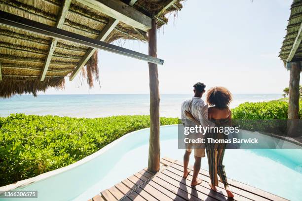 wide shot rear view of couple standing poolside at luxury tropical beachfront villa looking at view - over the shoulder view stockfoto's en -beelden