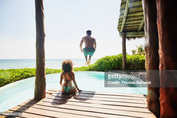 Wide shot rear view of man jumping into pool at luxury tropical villa while girlfriend sits poolside