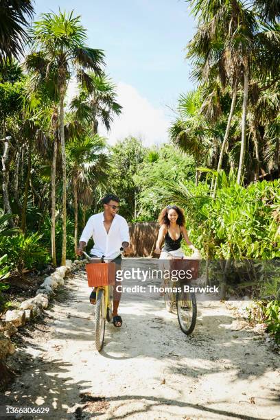 wide shot of smiling couple riding bikes to beach at tropical resort - tropical climate stock-fotos und bilder