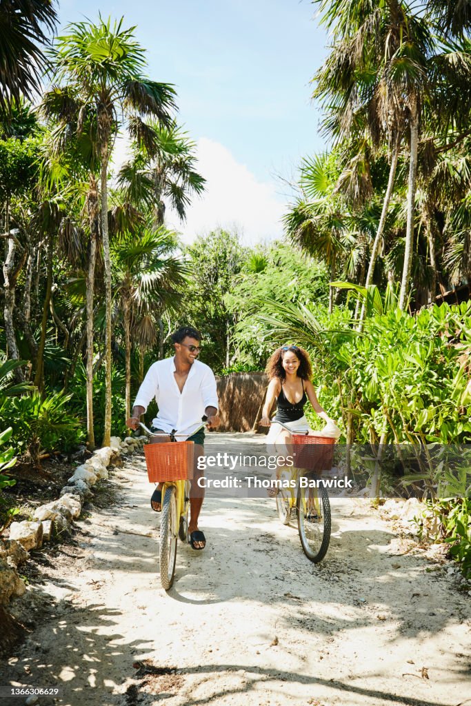 Wide shot of smiling couple riding bikes to beach at tropical resort
