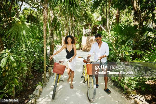 wide shot of smiling couple walking bikes on pathway in jungle at tropical resort - beach mexico bildbanksfoton och bilder