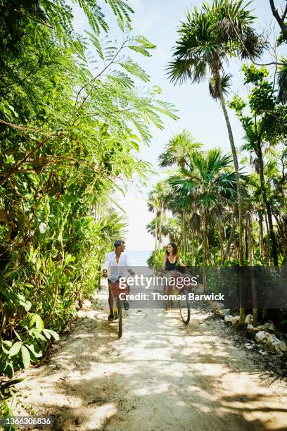 wide shot of smiling couple riding bikes from beach at tropical resort - mexico black and white stock-fotos und bilder