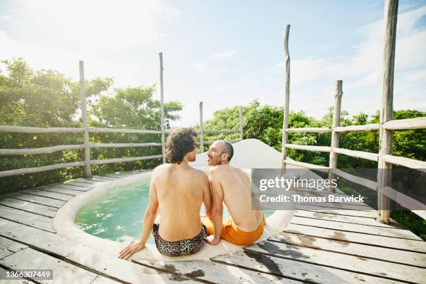 wide shot rear view of smiling gay couple sitting on edge of rooftop pool at eco resort in jungle - rooftop pool stock-fotos und bilder