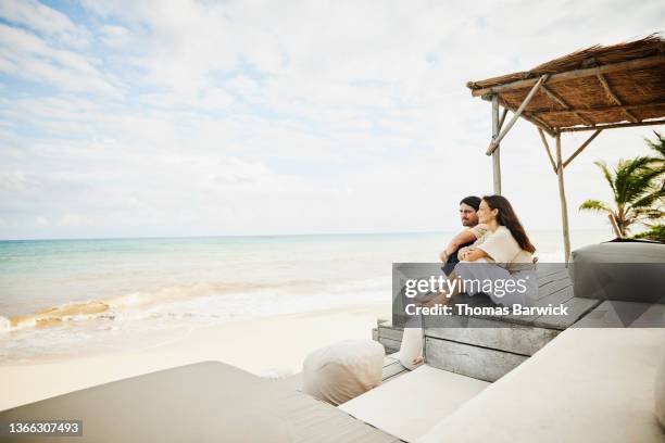 wide shot of smiling couple relaxing in tropical resort lounge area overlooking beach - kustegenskap bildbanksfoton och bilder