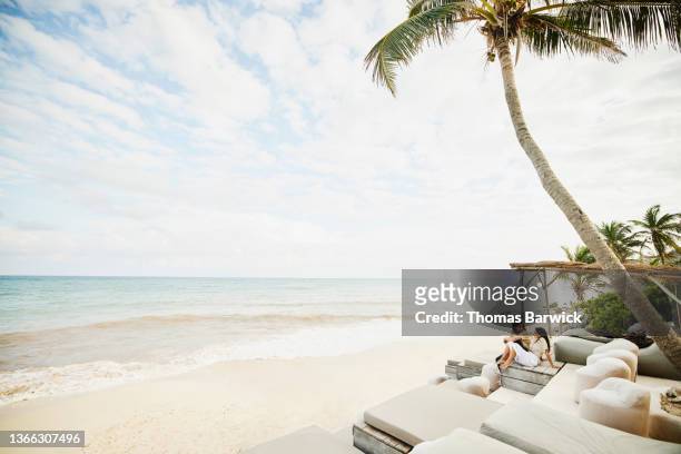 Extreme wide shot of smiling couple relaxing in tropical resort lounge area overlooking beach
