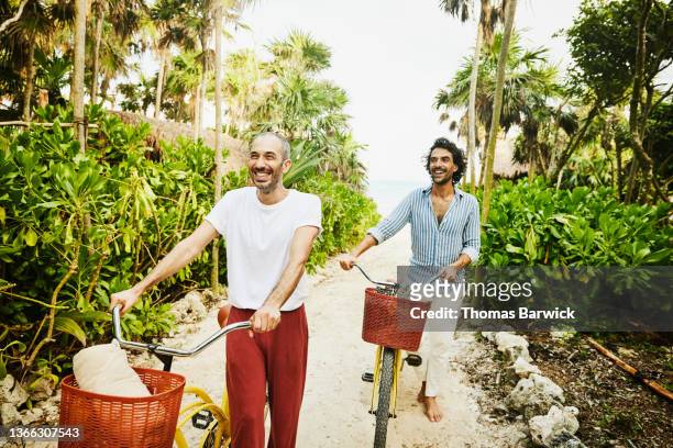 medium wide shot of laughing gay couple walking bikes on path from beach at tropical resort - handsome mexican men - fotografias e filmes do acervo