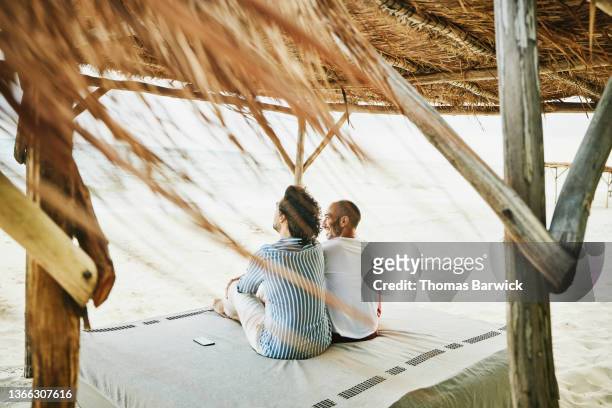 Wide shot rear view of gay couple relaxing under beach cabana at tropical resort