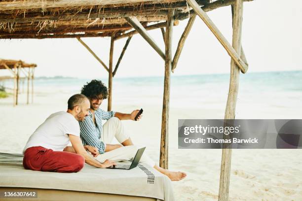 wide shot of smiling gay couple looking at data laptop while relaxing at beach cabana at tropical resort - travel copy space stock pictures, royalty-free photos & images