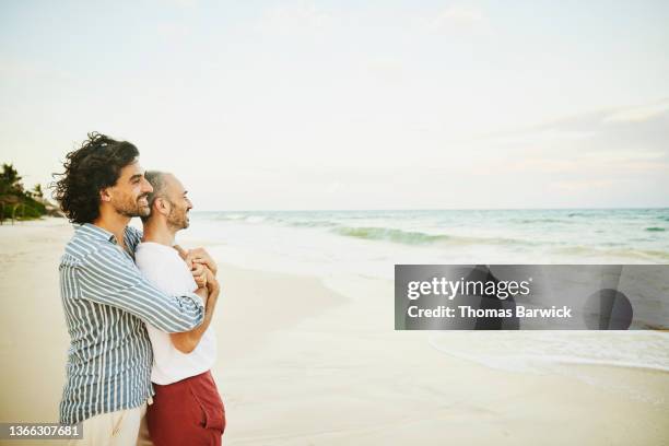 medium wide shot of embracing smiling gay couple standing on tropical beach looking at ocean - red dress shirt stock pictures, royalty-free photos & images