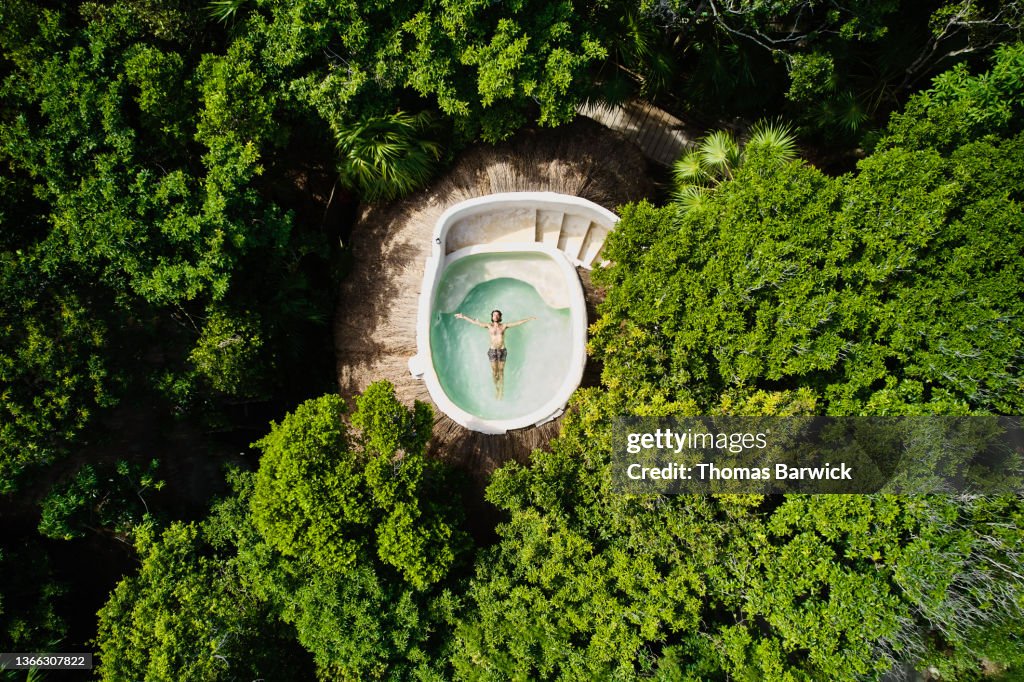 Wide shot overhead aerial view of man floating on back in rooftop pool at eco jungle resort