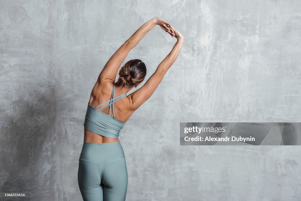 Woman doing back stretch practicing yoga standing against wall background with copy space.