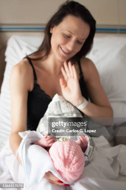 a young mother holds her newborn baby girl wearing a pink woolly hat and wrapped up in a blanket on her lap she she smiles and waves at her on a bed in a flat in edinburgh, scotland, uk - girls in bras photos stock pictures, royalty-free photos & images