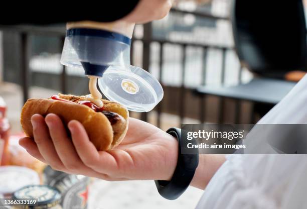close up of a person dressed in white holding a hot dog while putting mayonnaise sauce on it and some bars in the background. - mayonaise stockfoto's en -beelden
