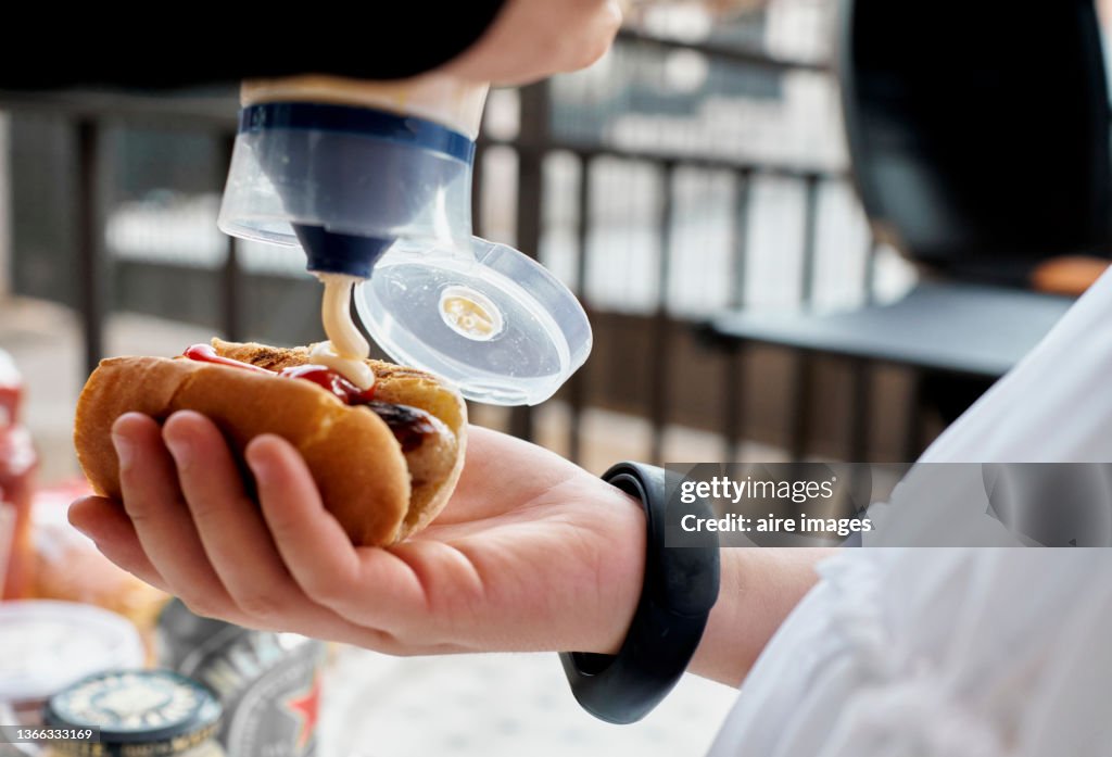 Close up of a person dressed in white holding a hot dog while putting mayonnaise sauce on it and some bars in the background.