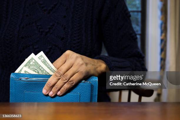 black woman holding vibrant blue wallet with multiple $1 u.s. paper bills showing while sitting at desk at home - pay cash stock pictures, royalty-free photos & images