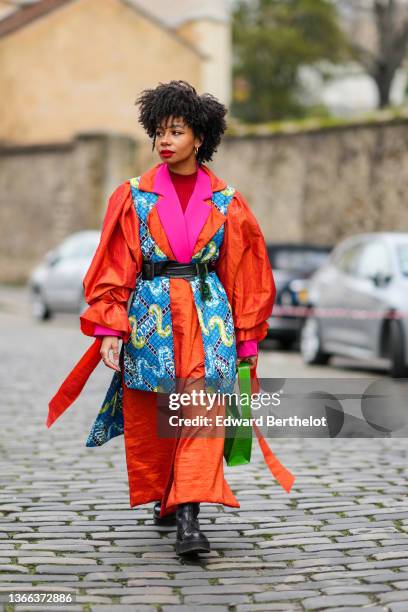 Sarah Monteil wears gold earrings, a red ribbed wool high neck pullover, a neon pink blazer jacket, an orange puffy sleeves long coat, a blue with...