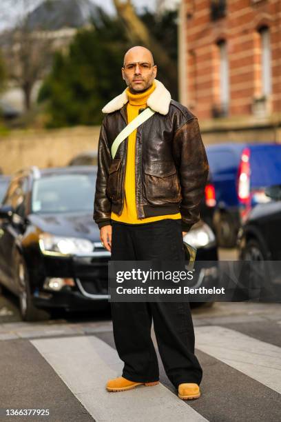 Mads Emil Grove Moller wears sunglasses, silver and gold earrings, a yellow wool ribbed turtleneck pullover, a brown shiny leather with white sheep...