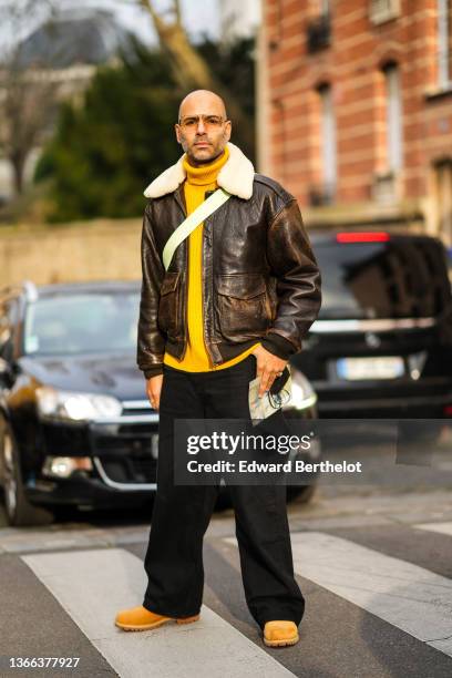 Mads Emil Grove Moller wears sunglasses, silver and gold earrings, a yellow wool ribbed turtleneck pullover, a brown shiny leather with white sheep...