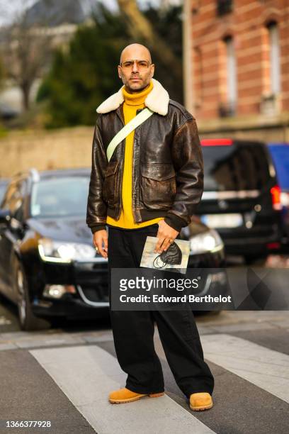 Mads Emil Grove Moller wears sunglasses, silver and gold earrings, a yellow wool ribbed turtleneck pullover, a brown shiny leather with white sheep...