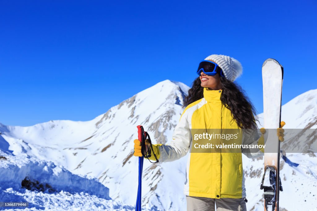 Frau mit gelber Jacke, die lächelt und ihren Winterurlaub genießt, mit Skiausrüstung in einem Berghintergrund mit strahlend blauem Himmel