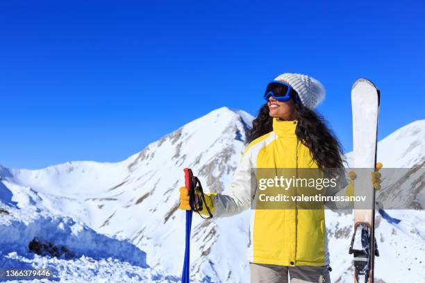 woman with yellow jacket smiling and enjoyng her winter vacation, holding ski equipment on a mountain background with bright blue sky - ski jacket stock pictures, royalty-free photos & images