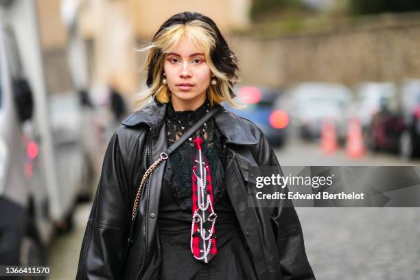 Guest wears gold and pearls pendant earrings, a black lace print pattern shirt, a black and red checkered print pattern zipper / belted tie, a black...