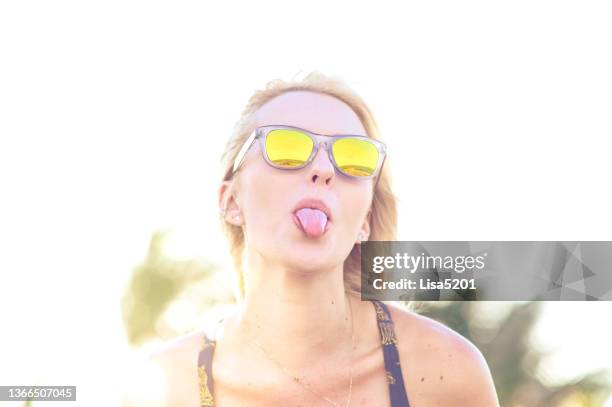 beautiful quirky young woman in sunglasses sticking out her tongue to be playful - cheesy grin stock pictures, royalty-free photos & images