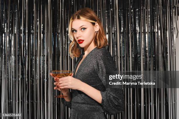 the girl stands smiling and holds a glass in her hands. tinsel silver. red lipstick. hollywood style. corporate party. portrait of a beautiful smiling blonde. the girl at the new year's party. new year party. curly hair. silver dress. - curly red hair glasses stock pictures, royalty-free photos & images