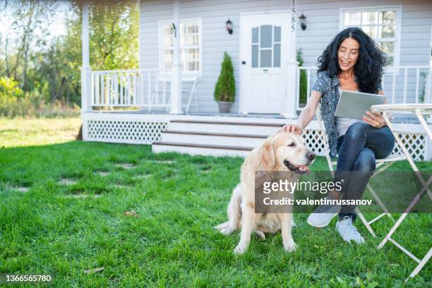 curly women sitting in front of the porch of her house, enjoying sunny day, holding digital tablet and playing with her retriever dog. - the house of flaunt oscar retreat hosted by manuel day 1 stockfoto's en -beelden