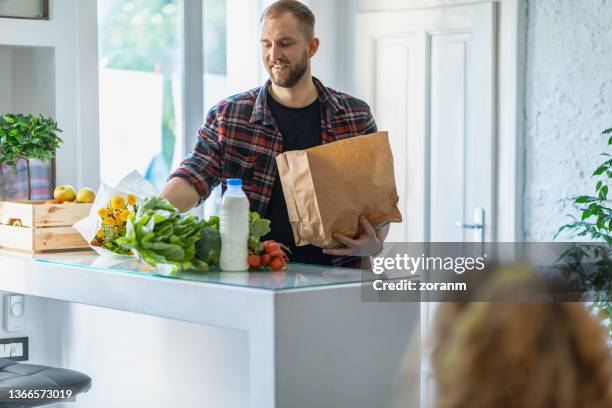happy young man taking groceries out of paper bag on the counter at home for making lunch - young man groceries kitchen stock pictures, royalty-free photos & images