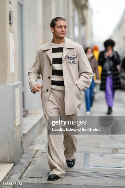 Gabriele Esposito wears silver and gold earrings, a black and white striped wool pullover, a beige long coat with a navy blue embroidered...