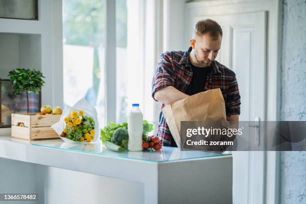 young man taking groceries out of paper bag on the counter at home for making lunch - young man groceries kitchen stockfoto's en -beelden